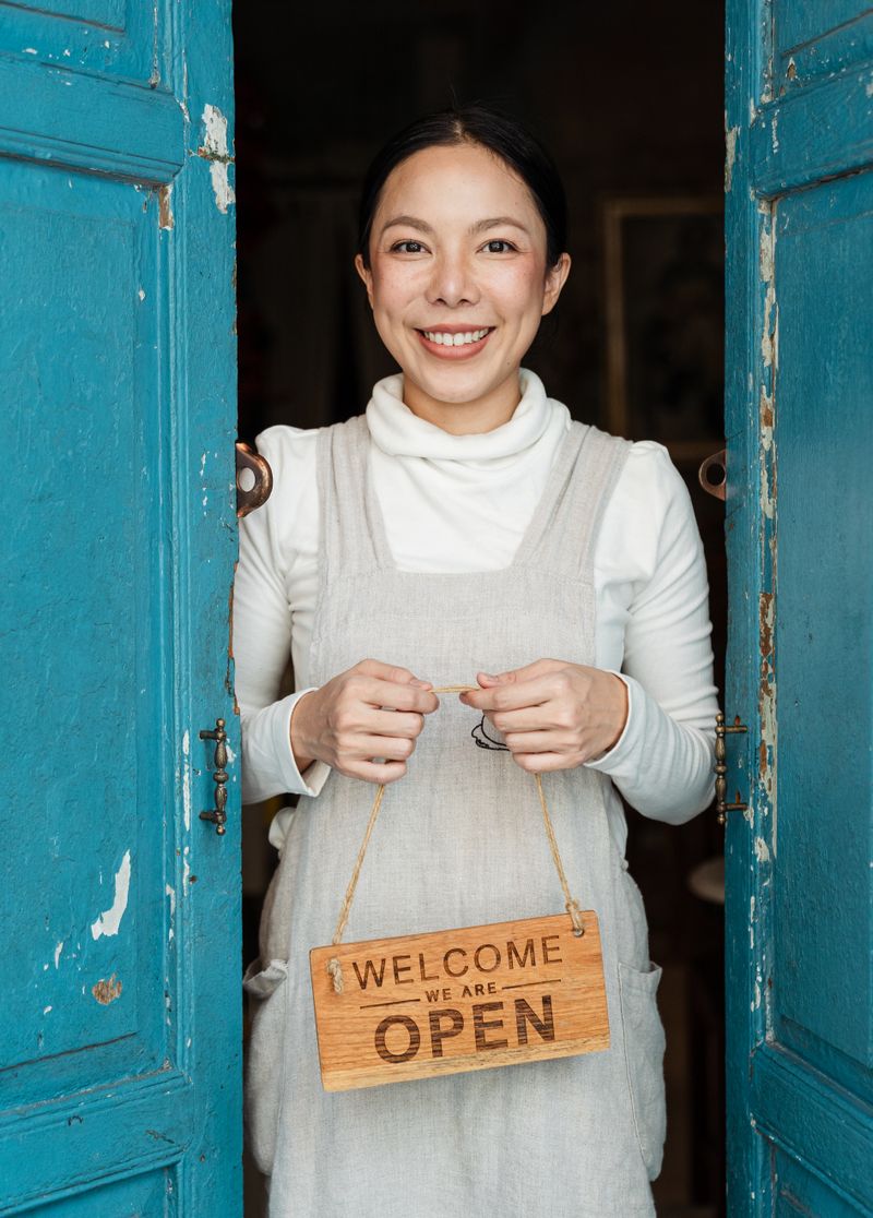 A smiling shop owner in an apron stands in her doorway holding a wooden sign that says Welcome, we are open.